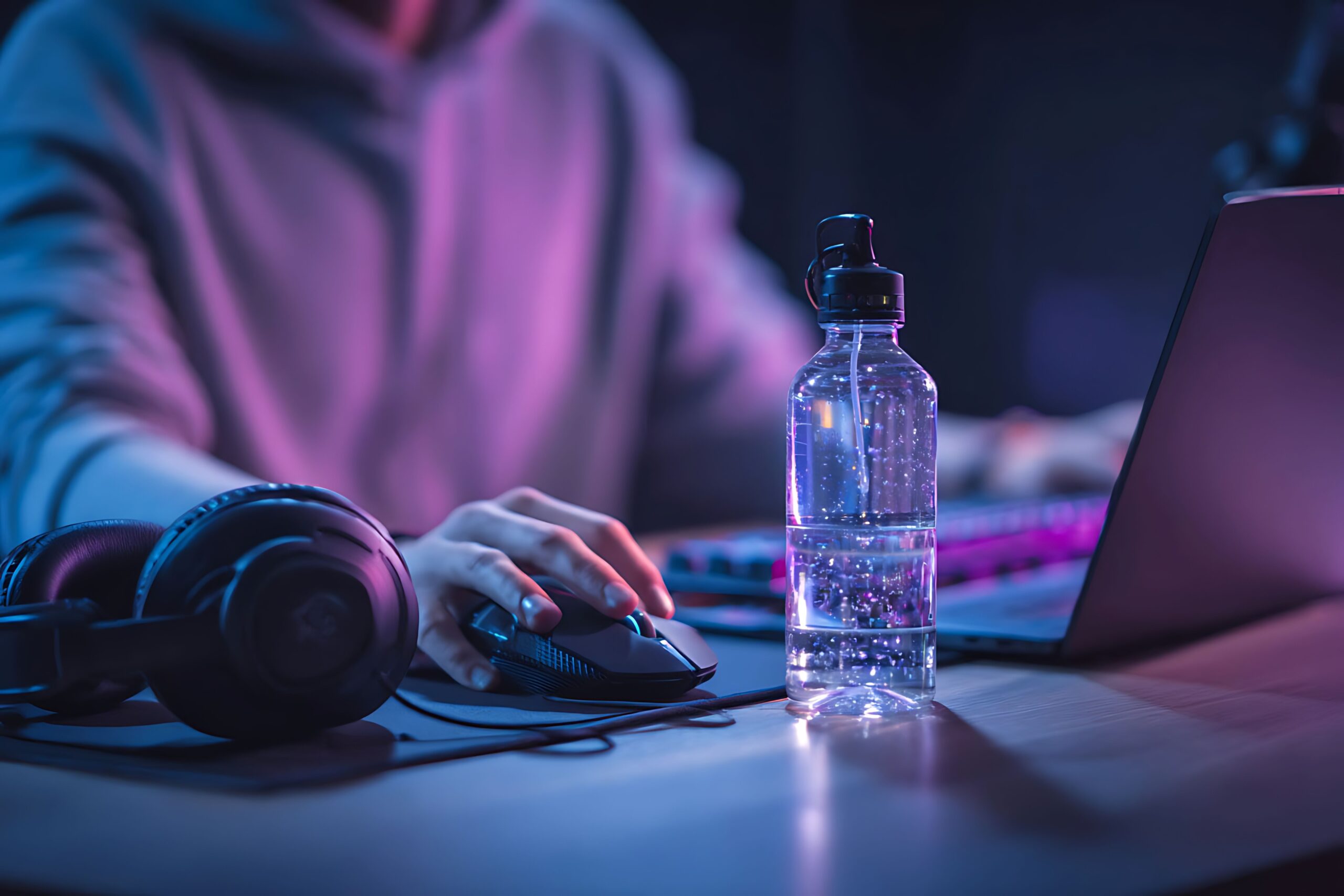 Rent a minecraft server: A person in a hoodie is using a gaming mouse and laptop in a dimly lit room with purple and blue ambient lighting